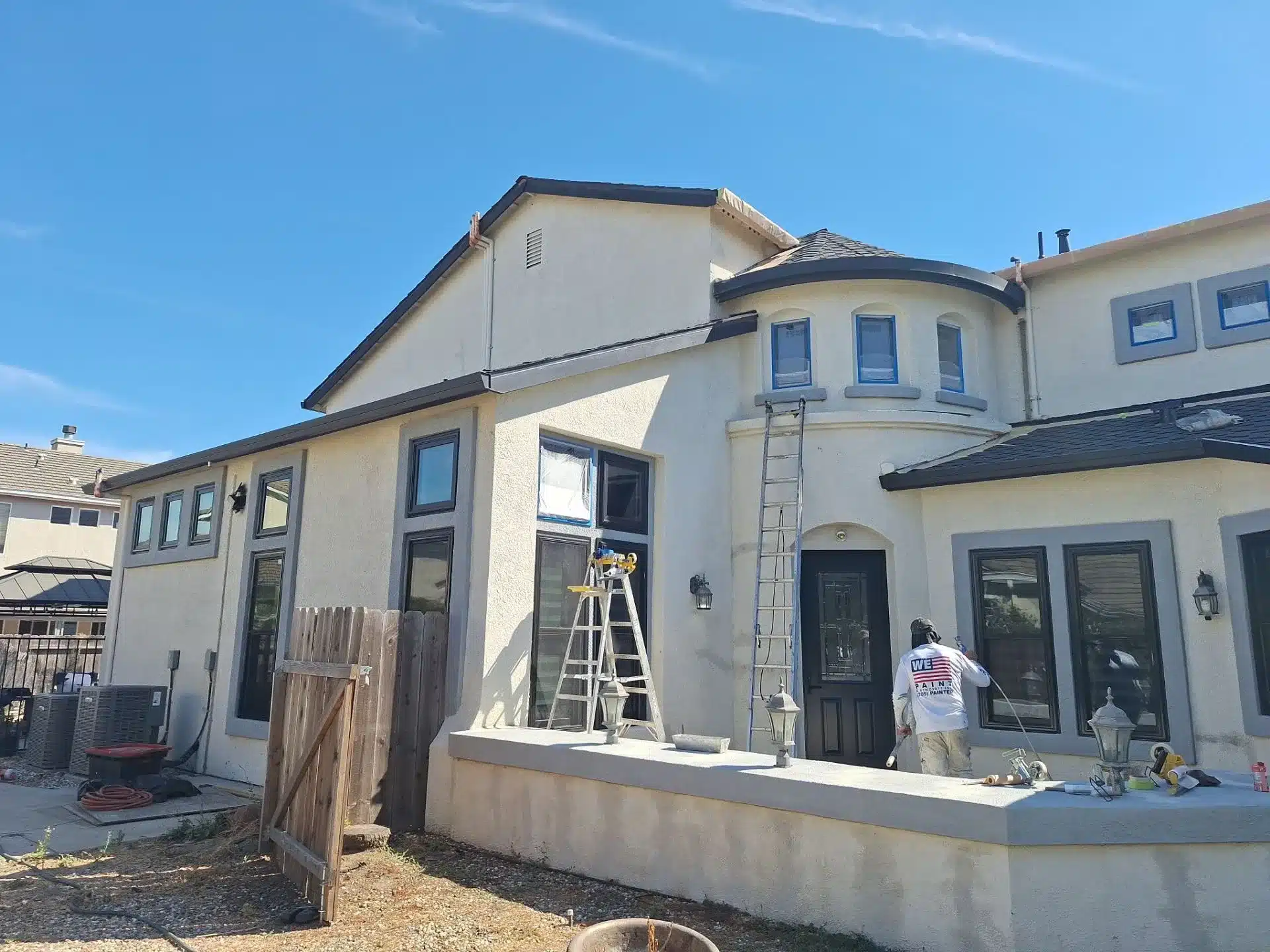Exterior Residential Home Painting Cream stucco residential home exterior with dark roof, multiple windows, and painter on ladder during exterior painting